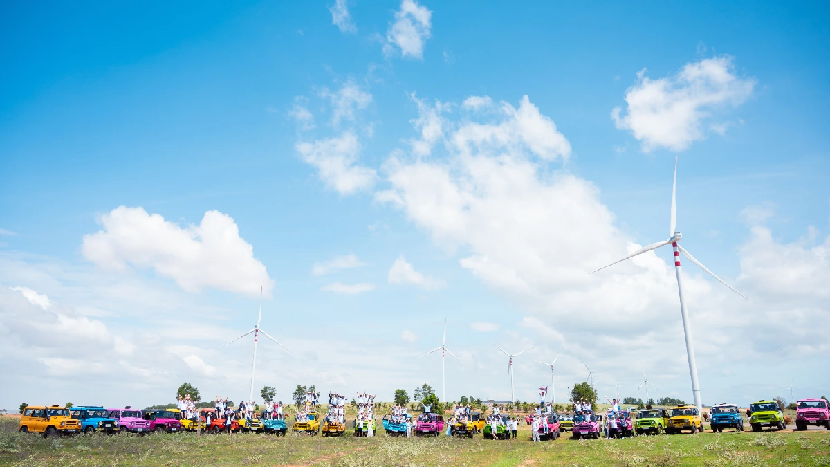 🖼️ Alt Text Colorful off-road vehicle convoy and a large group of people posing in a field with white wind turbines under a bright blue, cloudy sky