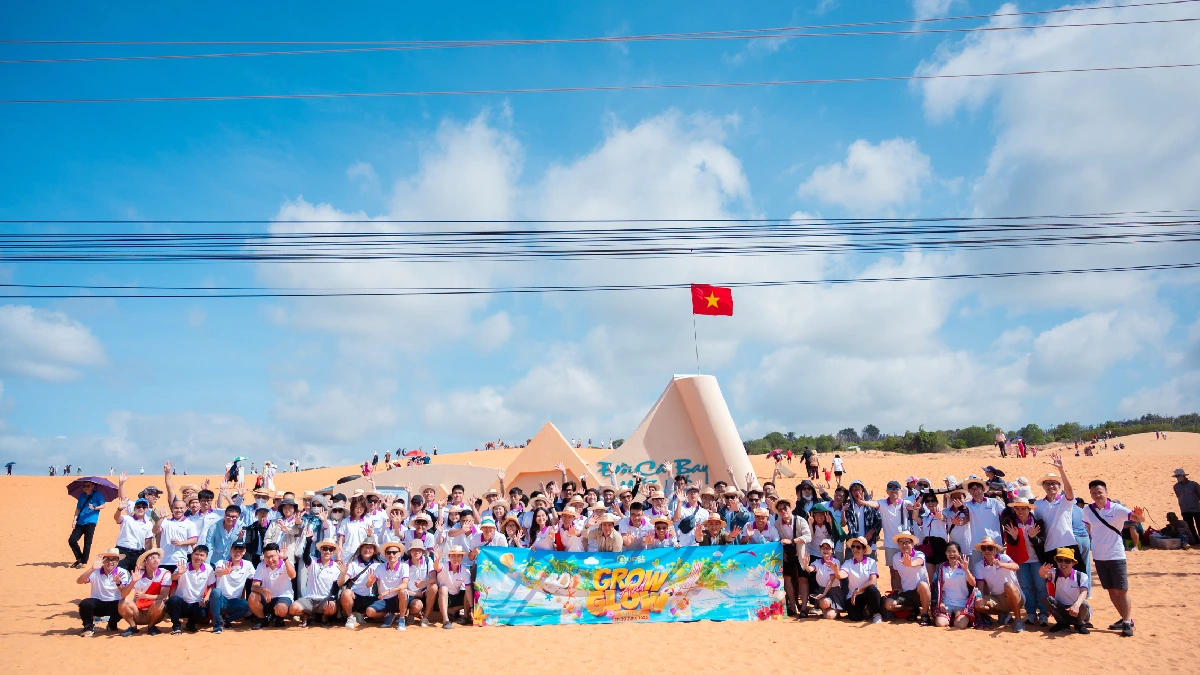 Large group posing with a banner on a sandy dune under a blue sky, with the Vietnamese flag flying from a background structure